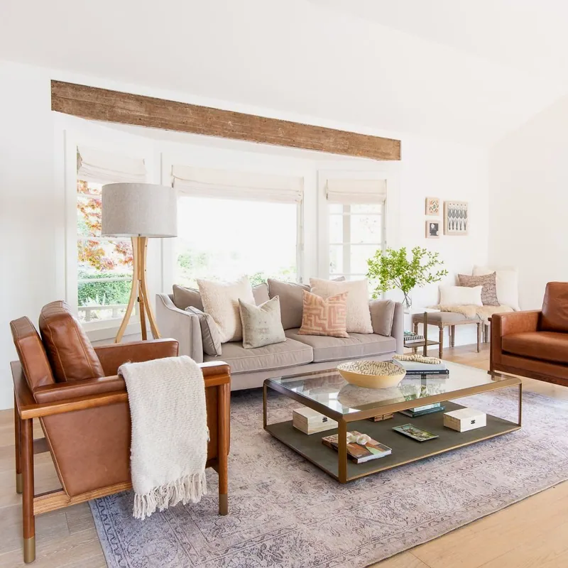 Bright living room with a beige sofa, leather armchair, coffee table, and decorative pillows, with large windows and wooden accents.