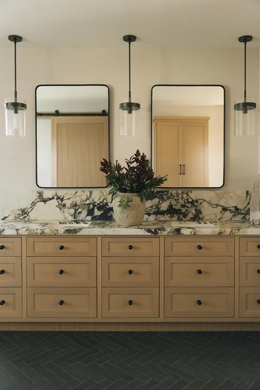 Bathroom vanity with light wood cabinets, marble countertop, two rectangular mirrors, a potted plant, and pendant lighting fixtures.