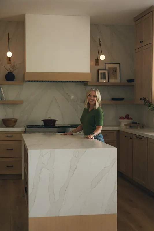 Designer Meredith Gough in a green dress beside a light-wood kitchen island with a custom range hood and marble counters.
