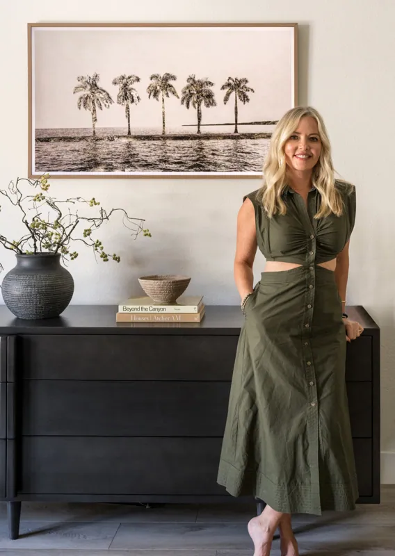 Meredith Gough in an olive dress in a dark wood-paneled room, styling a console with vases and framed botanical art.