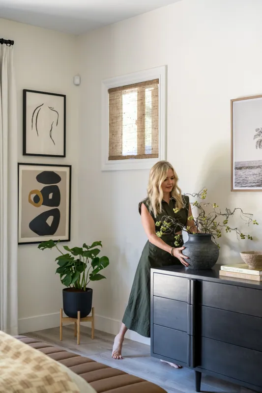 An interior designer with blonde hair, wearing a dark green dress, arranging and styling large black vase with branches on a black sideboard in a well-decorated living room which could also be a primary bedroom.
