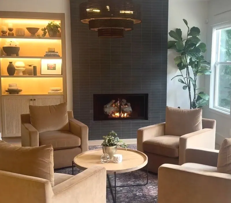 Living room with a fireplace, two cream armchairs, a round coffee table, and warmly lit built-in shelving.