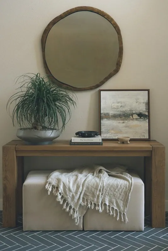 Entry vignette with a wood console, round mirror, framed art, a trailing plant, and a draped throw against a textured wall.
