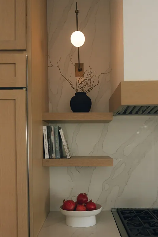 A kitchen with wooden shelves, a black vase with branches, a white bowl with pomegranates, a shelf with cookbooks, and a wall-mounted light fixture.