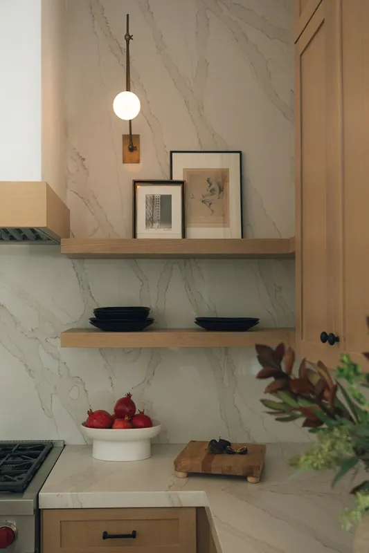 A kitchen corner with a marble backsplash, a wooden shelf with framed artwork, black bowls, pomegranate in a white bowl, cutting board, and a vase with greenery.