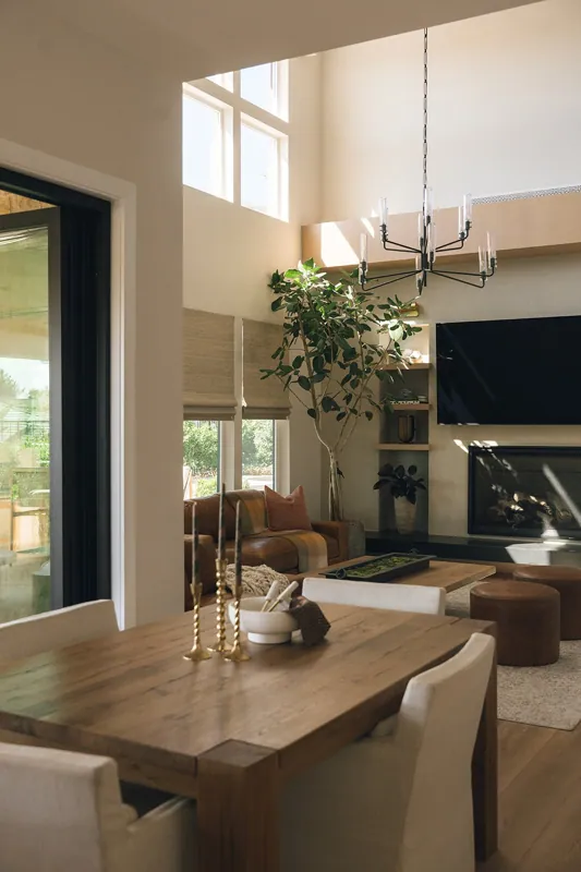 A living room with high ceilings, large windows, a modern chandelier, a wooden dining table with candles, a leather couch with cushions, a tall indoor plant, a television, and a fireplace.
