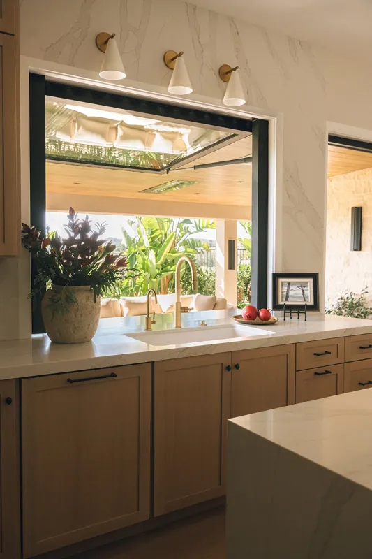 A kitchen with a large window showing a patio with greenery outside, beige cabinets, a white marble countertop, a gold faucet, red apples on a plate, a potted plant, and a picture frame on the counter.