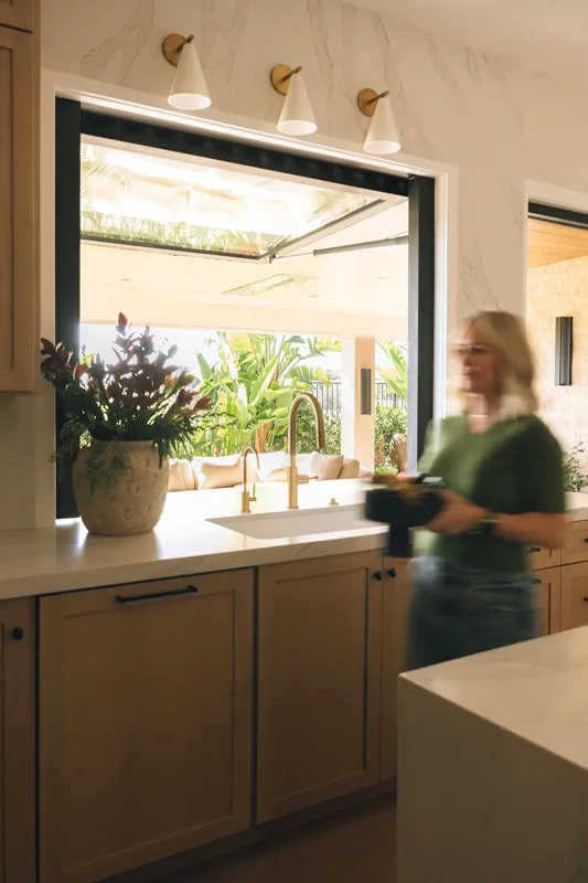A woman in motion holding a camera in a modern kitchen with a window looking out to a lush outdoor patio with greenery and outdoor seating.