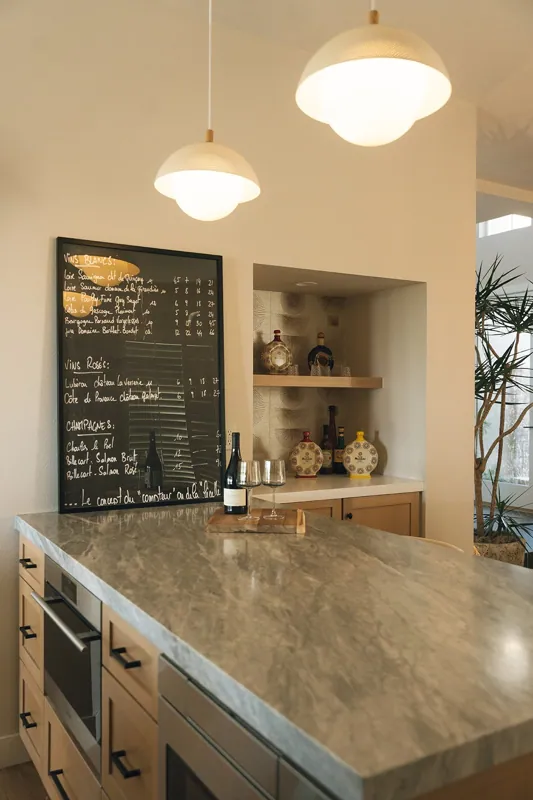 A modern kitchen with two hanging pendant lights, a marble countertop, and a chalkboard menu with handwritten wine and champagne options. There are wine bottles and glasses on the counter and decorative vases on a shelf in the background.