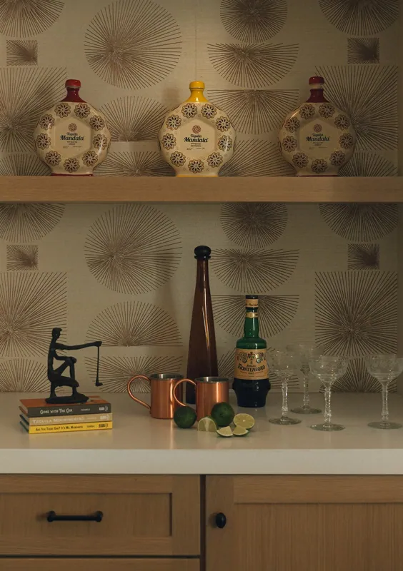 Decorative collection of spirits and barware on a kitchen countertop, featuring tequila bottles, a bottle of Montenegro, lime wedges, copper mugs, drinking glasses, and a small statue, with patterned wallpaper in the background.