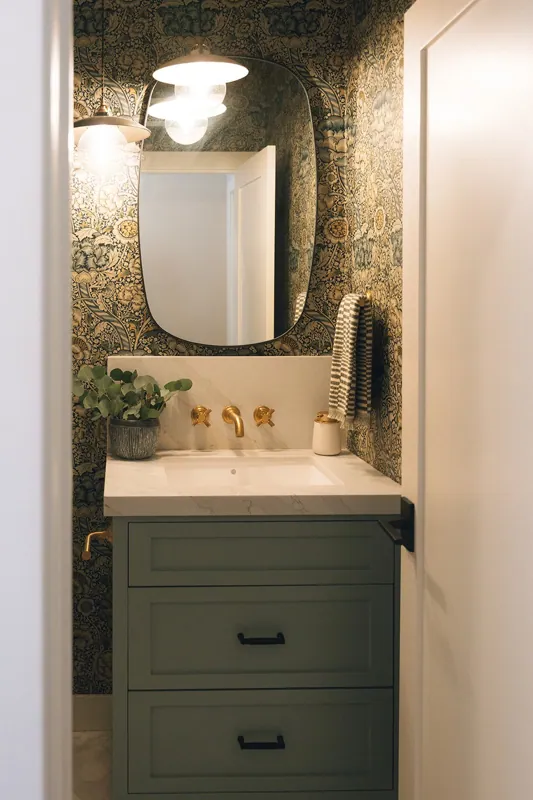 A small bathroom with a green vanity, gold fixtures, a plant in a gray pot, a striped towel, and a large irregularly shaped mirror facing a patterned wallpaper with floral and foliage designs.