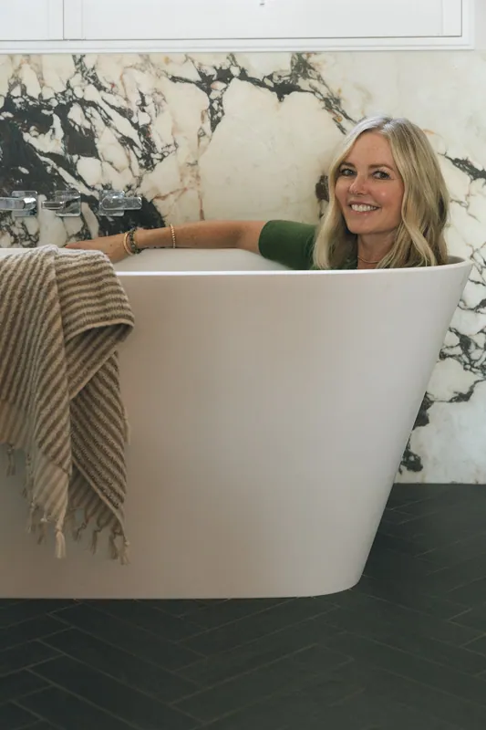 A woman with blonde hair and a green shirt sitting in a white bathtub, smiling at the camera, with a marble wall and a beige towel hanging over the side of the tub.