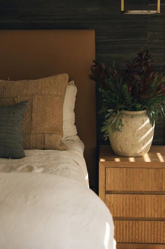 A bedroom with beige bedding, textured pillows, a wooden nightstand, and a large potted plant with purple and green leaves. The background includes a dark wood wall and a light fixture.