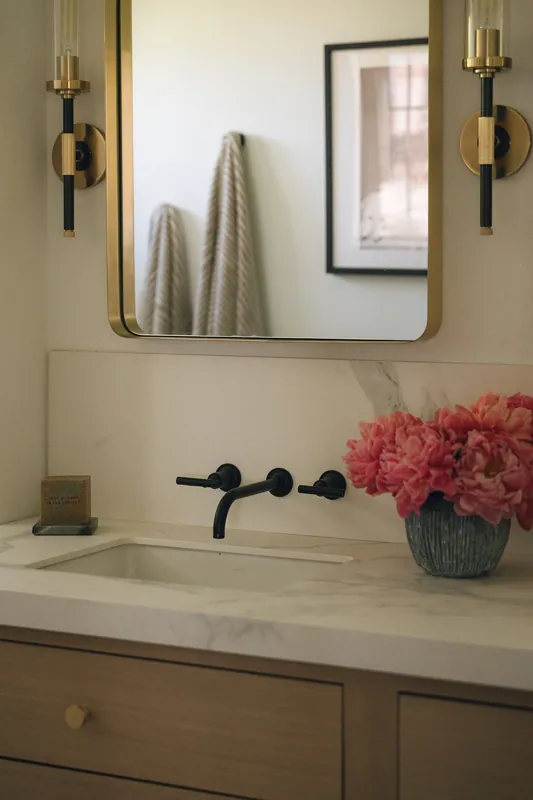 Bathroom vanity with a black faucet, pink flowers in a gray vase, and a white marble countertop, reflected in a mirror with gold and black sconces on either side.