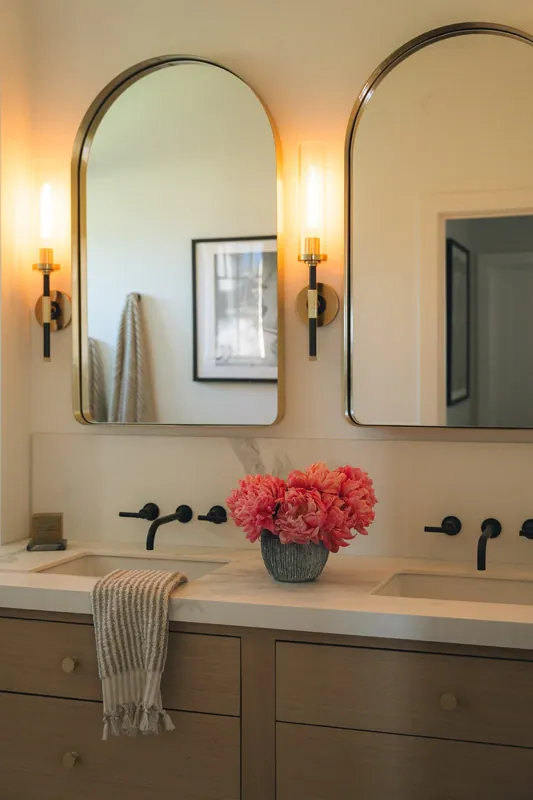 Bathroom double vanity with white countertop, two oval mirrors, two wall-mounted light fixtures, a vase with pink flowers, and a striped towel hanging over the sink.