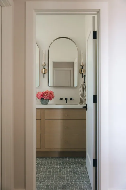 A bathroom seen through an open door with a wooden vanity, a white countertop, a pink flower arrangement, a large mirror, two light fixtures, and a towel hanging on the side.