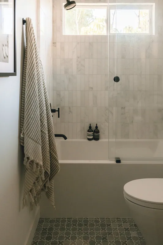 Modern bathroom with white marble tiles, a bathtub with two black soap dispensers, a glass shower door, a window with natural light, a white toilet, and a beige striped towel hanging on a black hook.