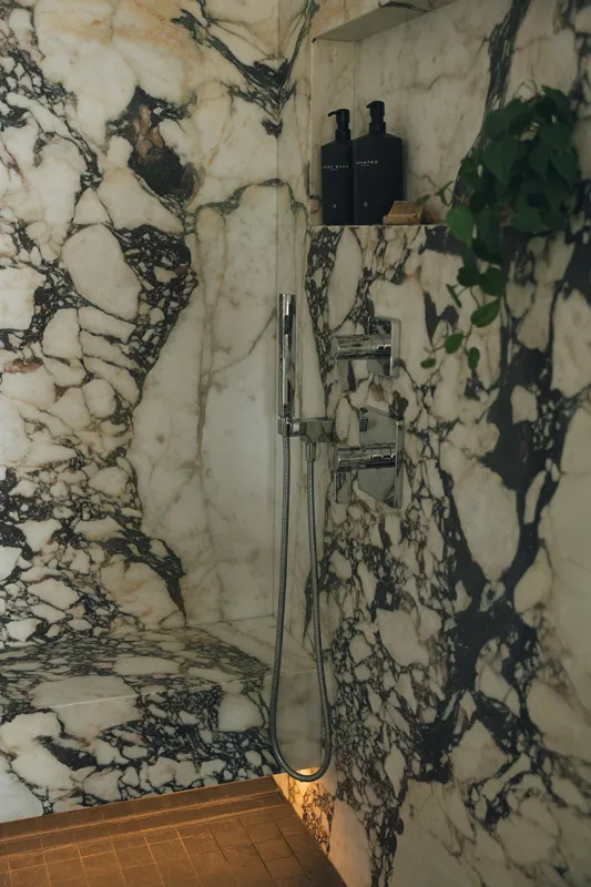 A marble shower with black and white streaks, a built-in shelf with black bottles, a metallic showerhead, and a handheld shower device. The floor is brown tiled with a small square pattern.