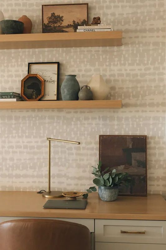 A home office or study space with a wooden desk, a closed laptop, a gold desk lamp, a woven coaster, a potted eucalyptus plant, and a framed mirror. Behind the desk are two floating shelves with decorative vases, framed pictures, books, and small sculptures.