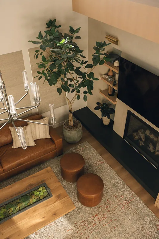 Living room with a brown leather sofa, beige pillows, a large potted plant, a black mantel with decor, a wall-mounted TV, a rug, and two round brown ottomans.