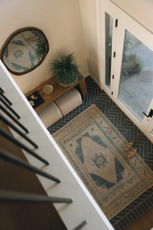 Top-down view of an entryway with a beige rug, a pair of beige shoes, a large green potted plant on a wooden bench, and a mirror on the wall. There is a glass door leading outside.