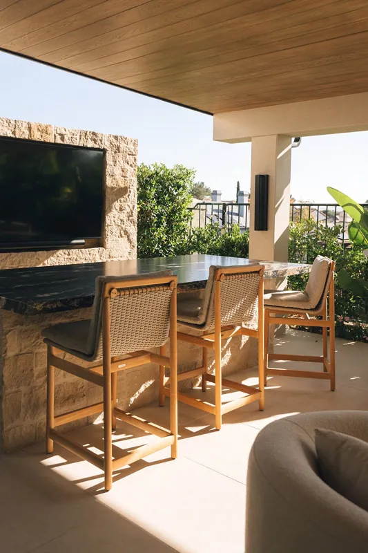 Outdoor patio with a black marble bar countertop, three beige cushioned wooden chairs, a stone fireplace with a TV, and lush green plants, with a view of a neighborhood in the background.