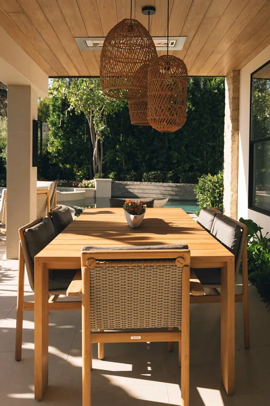 An outdoor dining table with six chairs, a flowerpot centerpiece, hanging wicker pendant lights, a wooden ceiling, and a view of lush green trees and pool in the background.