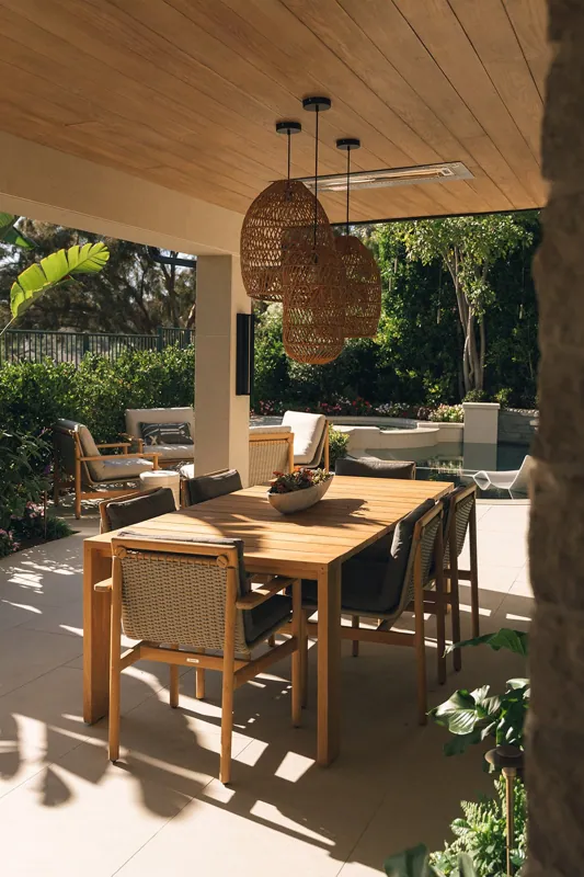 Outdoor patio with a wooden dining table and six chairs, hanging wicker pendant lights, and lush greenery and flowers in the background.