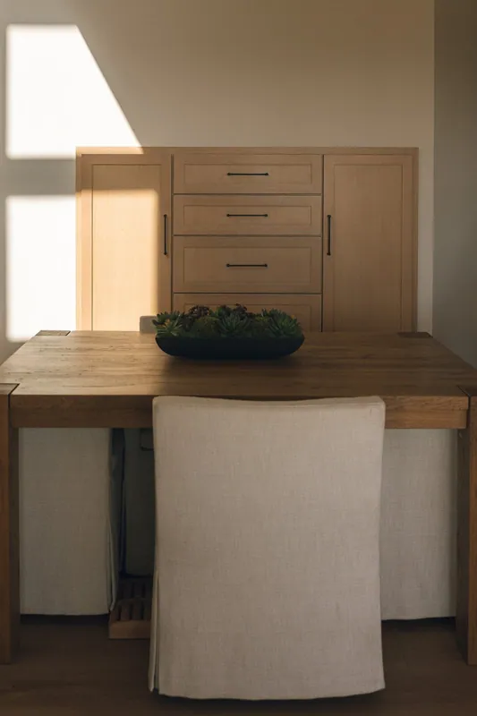 A wooden dining table with a beige upholstered chair facing a wooden cabinet with drawers. A black bowl with green succulents is on the table. Sunlight creates a shadow on the wall behind the cabinet.