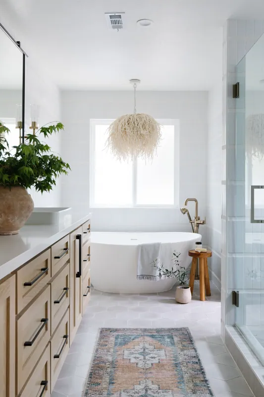 Modern bathroom with a white bathtub, wooden vanity, potted plants, a large window, and a decorative pendant light fixture.