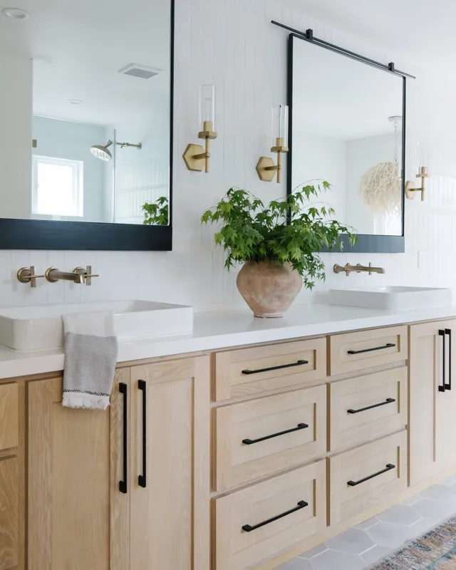 Bathroom double vanity with light wood cabinets, black handles, white countertop, two vessel sinks, large mirror, wall-mounted gold and black fixtures, plant in a terra cotta pot, wall sconces, and a shower with a window in the background.