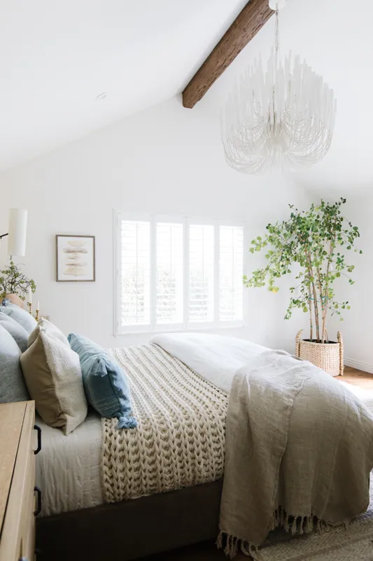 Bright bedroom with white walls, a large window with white shutters, a chandelier, a bed with neutral and blue pillows, a chunky knit throw, a beige blanket, a wooden nightstand, framed abstract artwork, and a large potted plant.