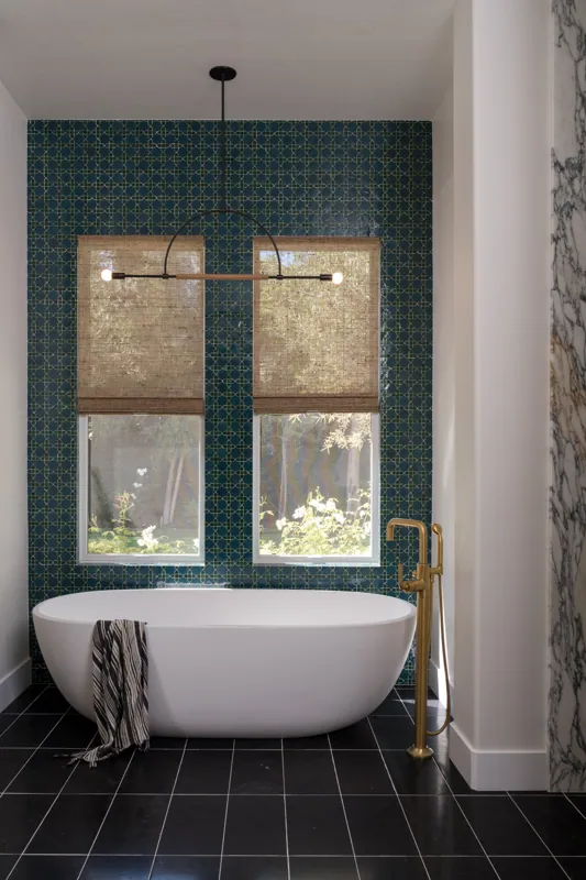 Modern bathroom with a freestanding bathtub in front of patterned wall with two windows. Floor has black tiles, and there is a gold floor-mounted faucet. A striped towel hangs over the bathtube.