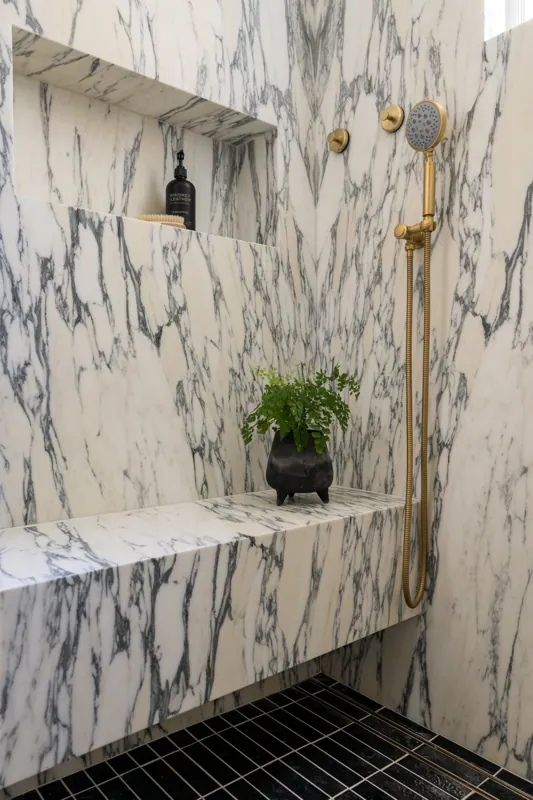A luxurious shower area with marble walls and a black tiled floor, featuring a brass showerhead, a small pot with green plants, and black bottles on the shelf.
