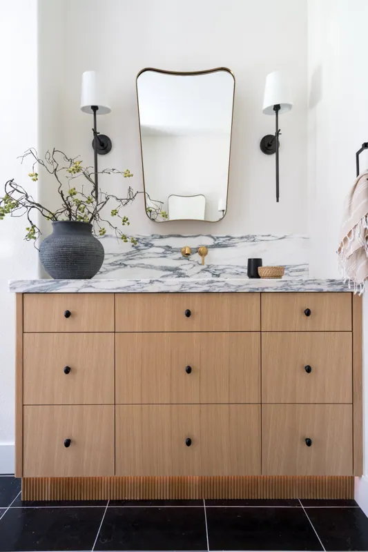 Bathroom vanity with a marble countertop, wooden cabinet, and black knobs. A narrow mirror with a wavy top, two wall sconces with white shades, a black vase with branches, and small decor items on the counter.