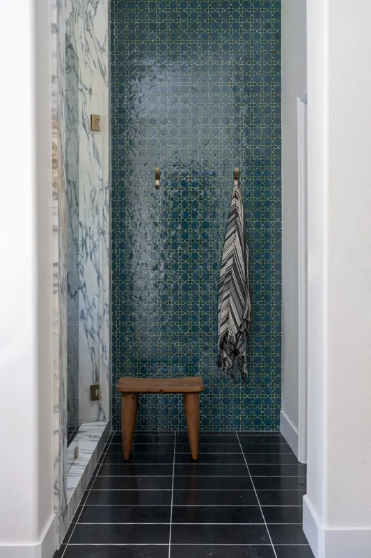A small tiled shower area with a dark teal geometric patterned wall, a marble wall to the left, and two brass hooks, one holding a striped towel, with a wooden stool in front.