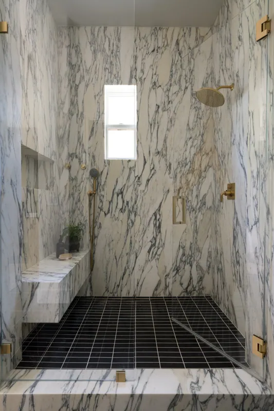 Luxurious marble shower with black tile floor, aged brass fixtures, small window, and built-in floating bench with plants. Tiled in drain. Luxury, low key, livable, casual California style primary shower.