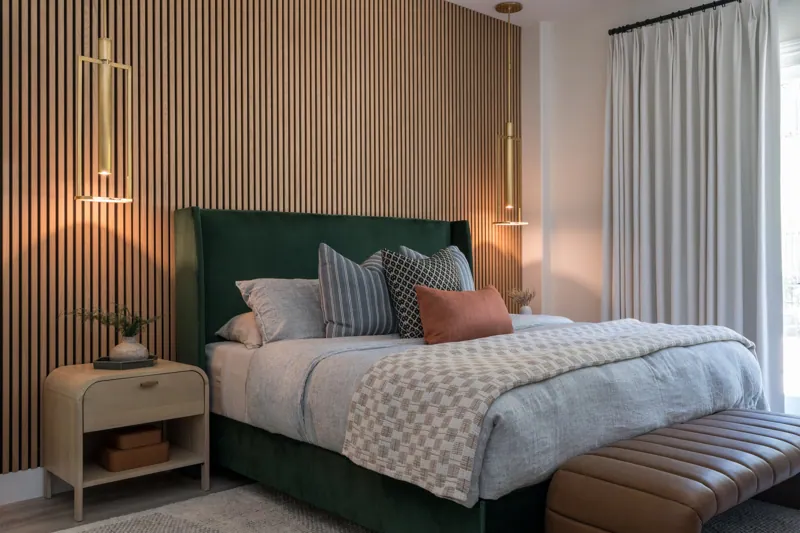 A modern bedroom with a green upholstered bed, gray bedding, patterned pillows, wooden accent wall, hanging pendant lights, bedside tables with vases and greenery, white curtains, and a leather bench at the foot of the bed.