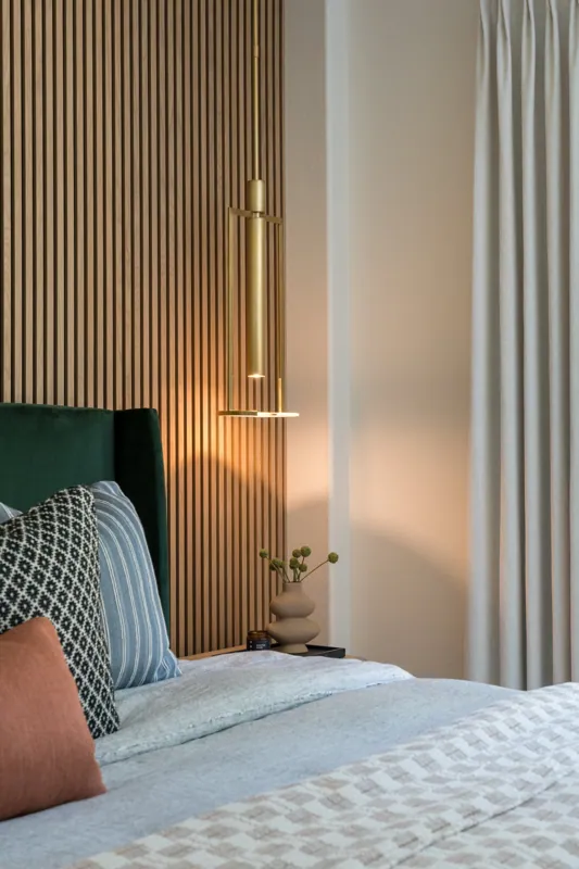 Bedroom with a green upholstered bed, patterned pillows, beige walls, vertical wood paneling, a modern gold pendant light, a vase with green round-topped flowers on a black nightstand, beige curtains.