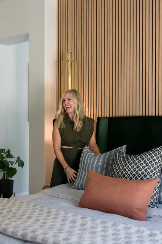 A happy interior designer with blonde hair wearing a green sleeveless dress, smiling and looking to the side in a modern bedroom with wooden wall paneling and decorative pillows on the bed.