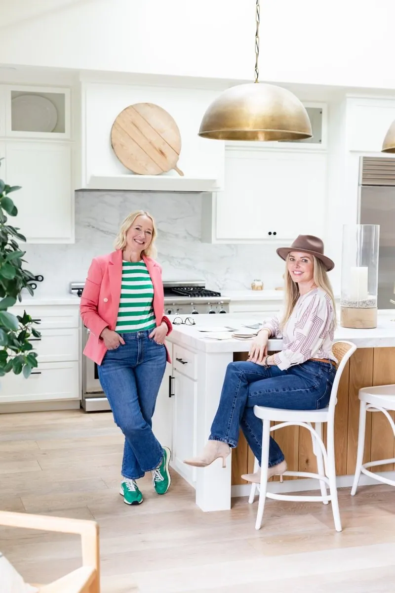 Two business women in a modern, bright kitchen with white cabinets and marble backsplash, one standing and one sitting on a bar stool with fabric samples on the countertop, smiling at the camera and preparing for a design materials meeting.