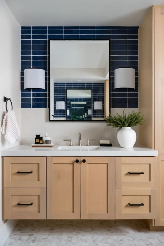 Bathroom vanity with light wood cabinets, a white countertop, a rectangular sink, a silver faucet, a large mirror with smaller mirrors behind it, blue wall tiles, two white wall sconces, a white vase with greenery, and bathroom toiletries.