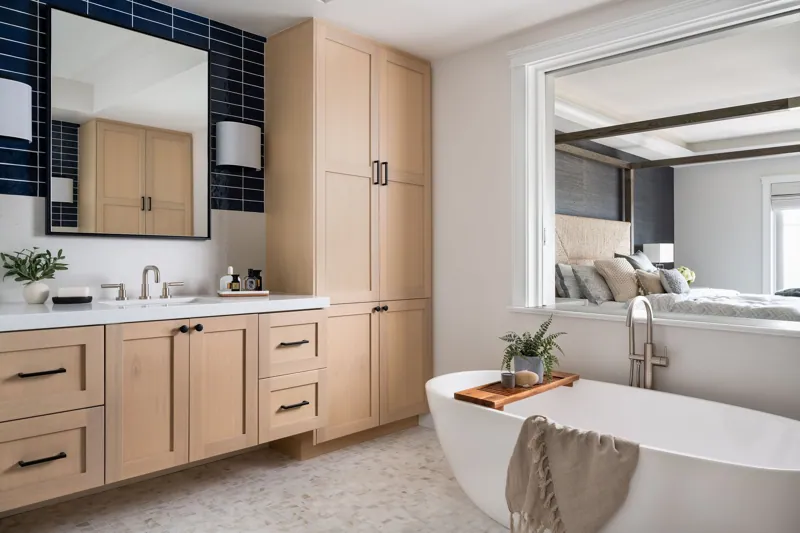 Modern bathroom with a white bathtub, beige cabinetry, a large mirror, and a separate adjacent bedroom visible through an open wall.