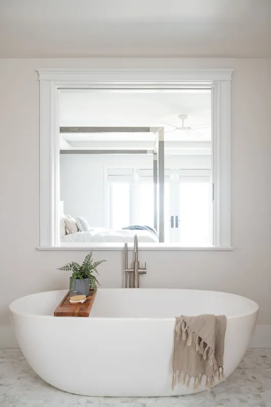 A modern bathroom with a white freestanding bathtub, a wooden tray with a potted plant and soap, a beige throw blanket, and a large window looking into a bedroom with a bed and ceiling fan.