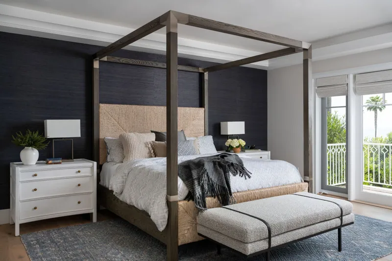 Bedroom with four-poster bed, white nightstands, two lamps, window with view of greenery and palm trees, and dark accent wall.