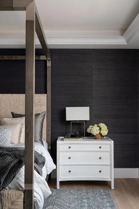 A bedroom with a bed on the left side, a white nightstand with a lamp, a vase of white flowers, and books on top, against a dark textured wall.