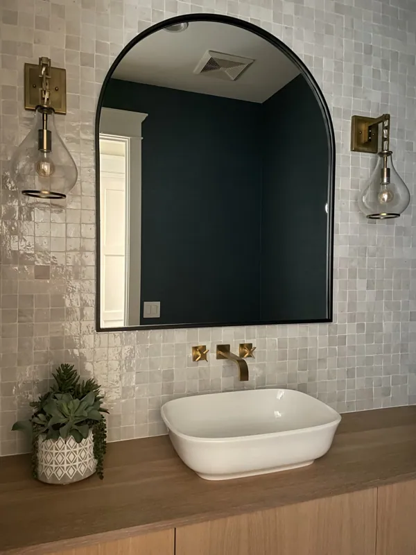 Bathroom vanity with a white vessel sink, gold faucet, two sconces with clear glass shades, large arched mirror, potted plant, wood countertop, and tiled wall.
