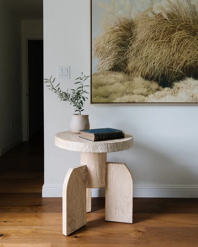 A wooden side table with a potted plant and a book on top, positioned against a white wall with a large painting of grasses hanging above. The floor is hardwood.