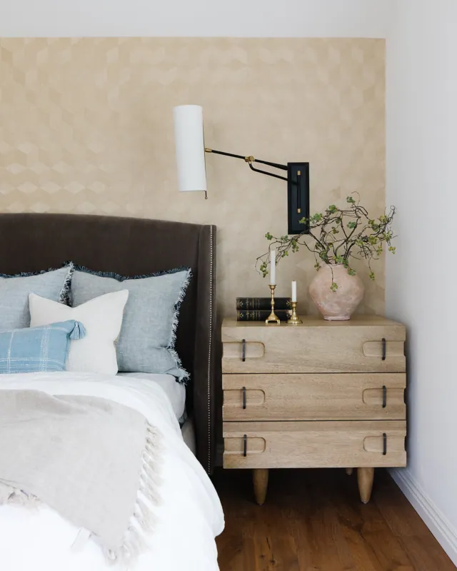 A bedroom with a bed that has multiple pillows, a dark headboard, and a wooden nightstand with a vase of branches, candles, and books. A wall-mounted reading lamp is above the nightstand and the wall has a geometric-patterned wallpaper.