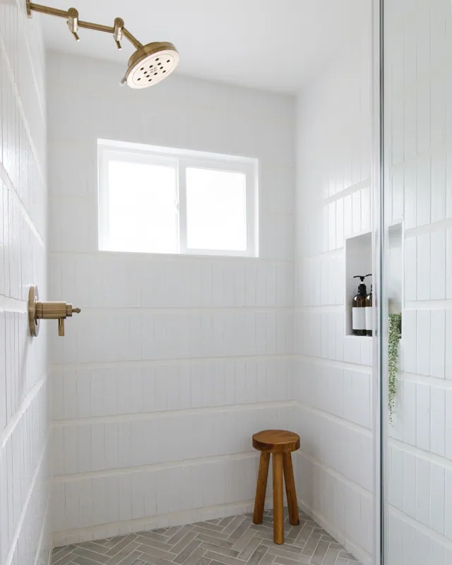 Minimalist white tiled shower with a window, a ceiling shower head, a small built-in shelf with soap, a wooden stool, and a glass shower door.